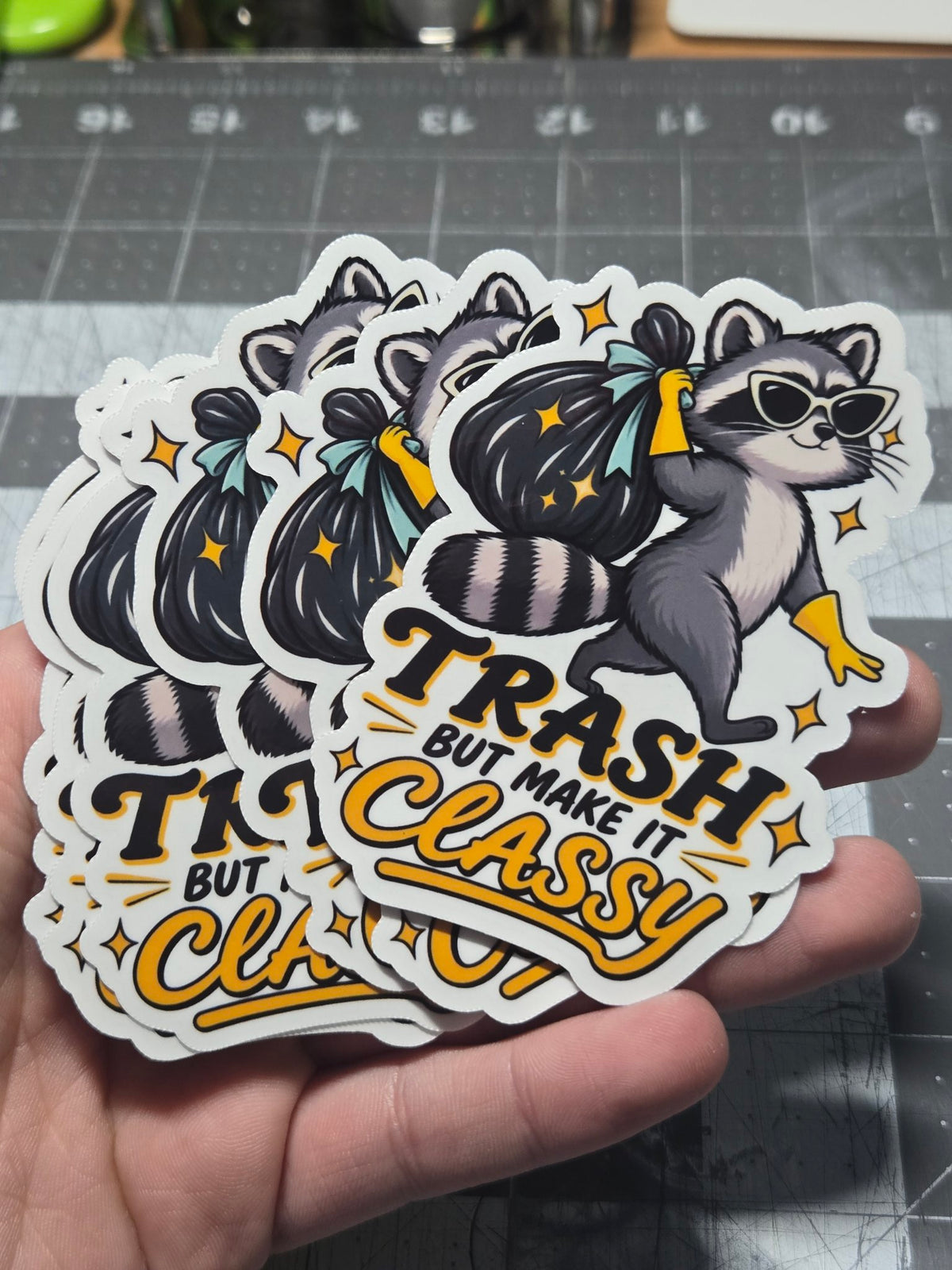 Hand holding "Trash but make it classy" raccoon vinyl stickers