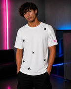 A man wearing a white short-sleeve t-shirt with black embroidered 'X' motifs and a pink embroidered logo on the sleeve, set against a dark studio background with pink and blue neon lights.