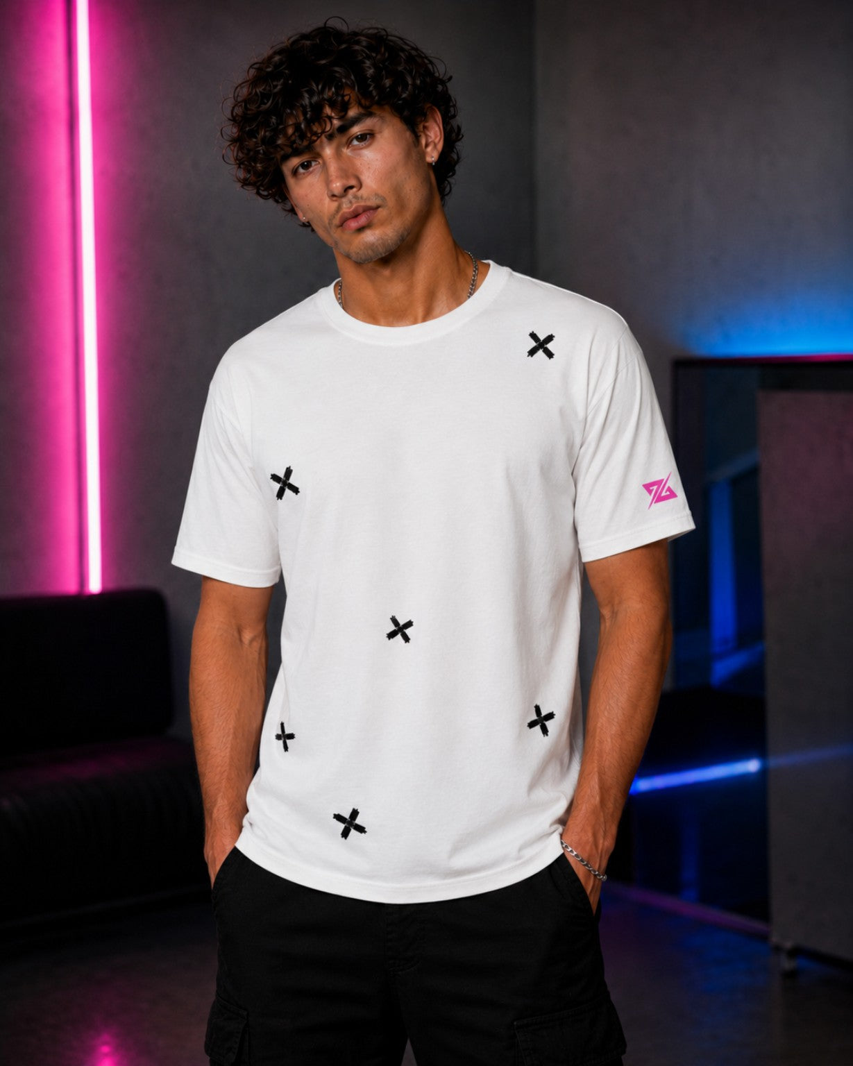 A man wearing a white short-sleeve t-shirt with black embroidered 'X' motifs and a pink embroidered logo on the sleeve, set against a dark studio background with pink and blue neon lights.